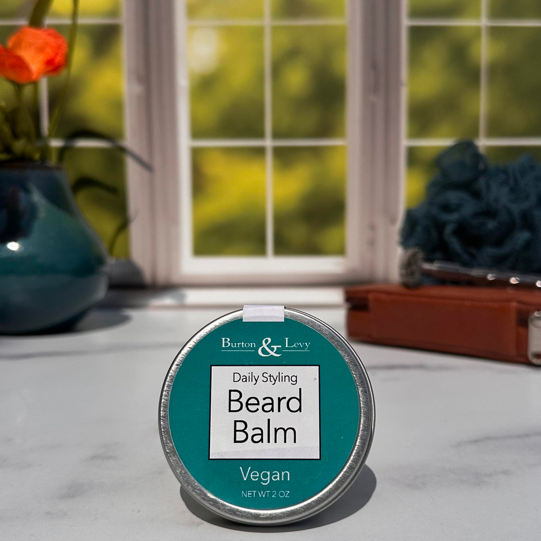 Burton & Levy Daily Styling Vegan Beard Balm in front of a window with a vase and razor off to the sides.
